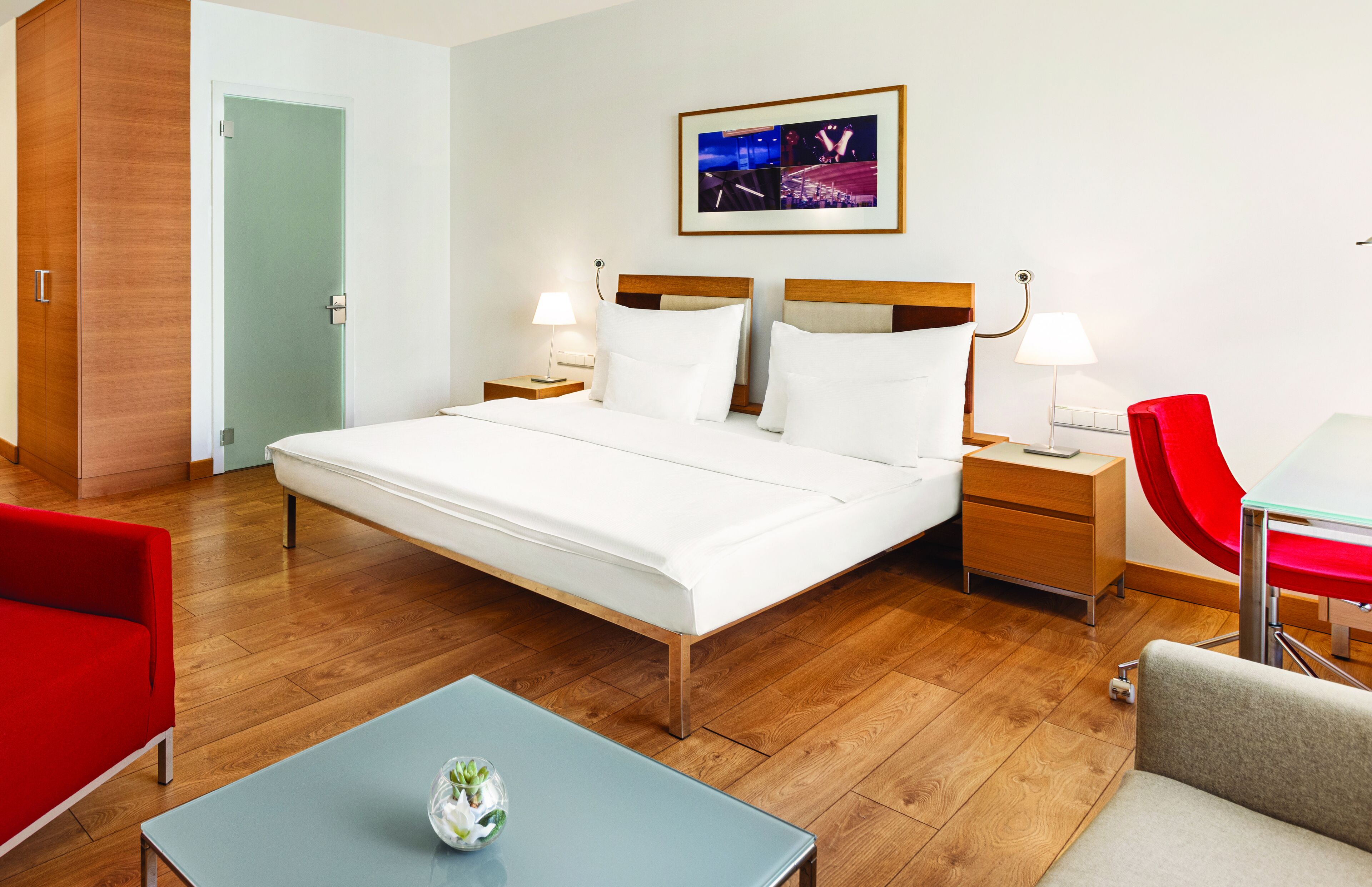 Une chambre d’hôtel avec un grand lit, deux tables de chevet avec des lampes, un fauteuil rouge, un canapé gris et une armoire en bois.