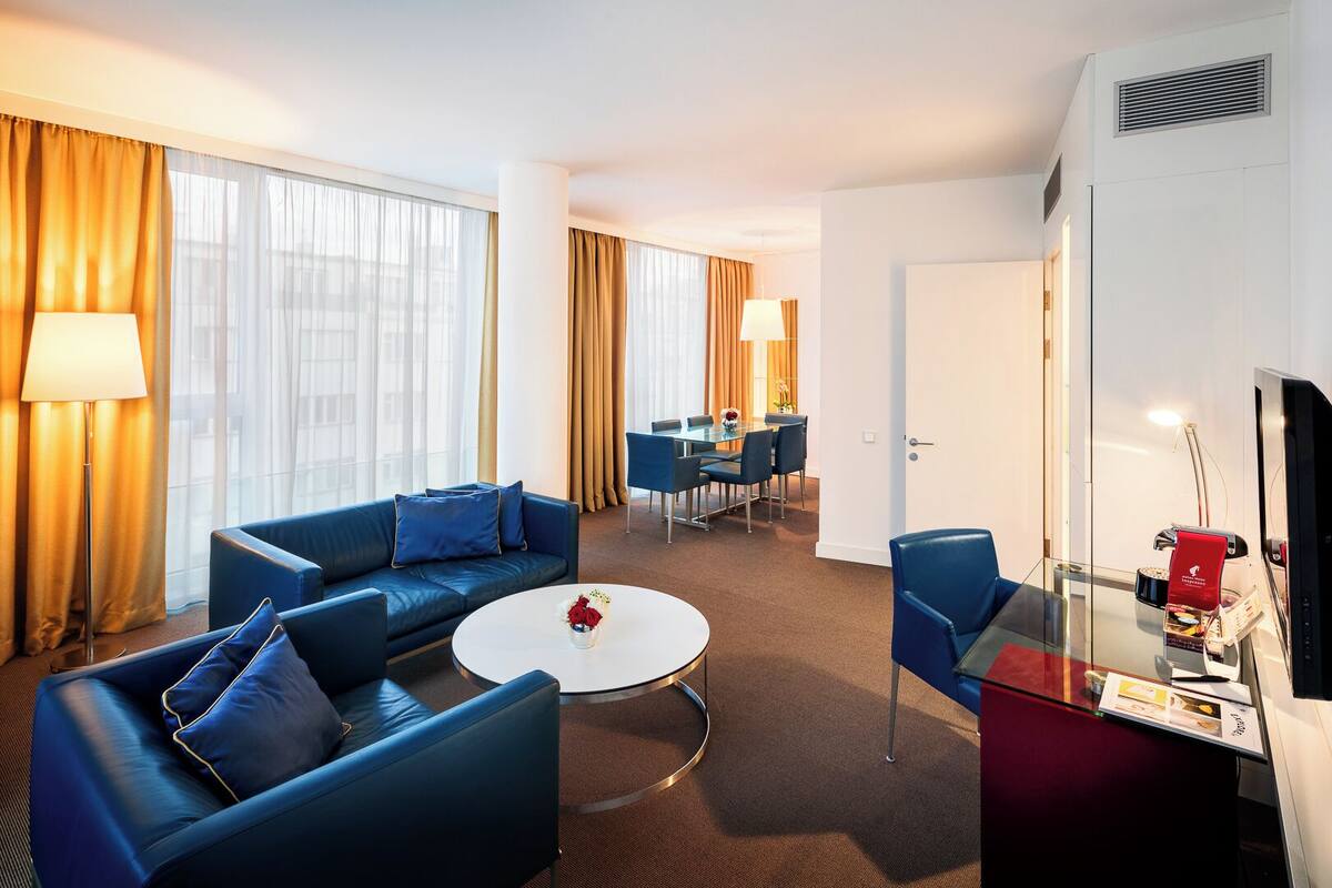 Ein modernes Hotelzimmer mit blauer Sofagarnitur, einem runden Couchtisch, einer Essecke mit Tisch und Stühlen sowie einem Fernseher auf einem Schrank.