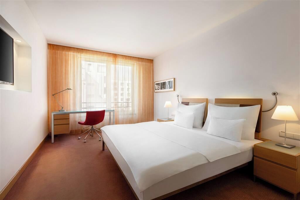 A hotel room with a large bed, a desk, a red chair, and a window with curtains.