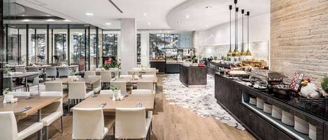 Un comedor moderno con mesas largas y sillas, una barra de buffet con diversas opciones de comida, y un amplio ventanal que ofrece vistas al exterior.