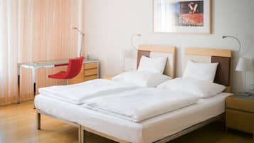 Uma cama de casal com lençóis brancos, uma escrivaninha com uma cadeira vermelha e uma mesa de cabeceira com abajur.