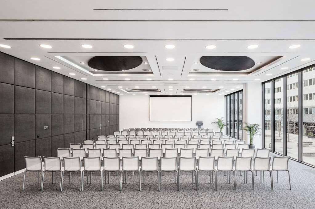 Un salón de conferencias con filas de sillas, una pantalla grande y vistas al exterior.