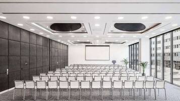 Un salón de conferencias con filas de sillas, una pantalla grande y vistas al exterior.