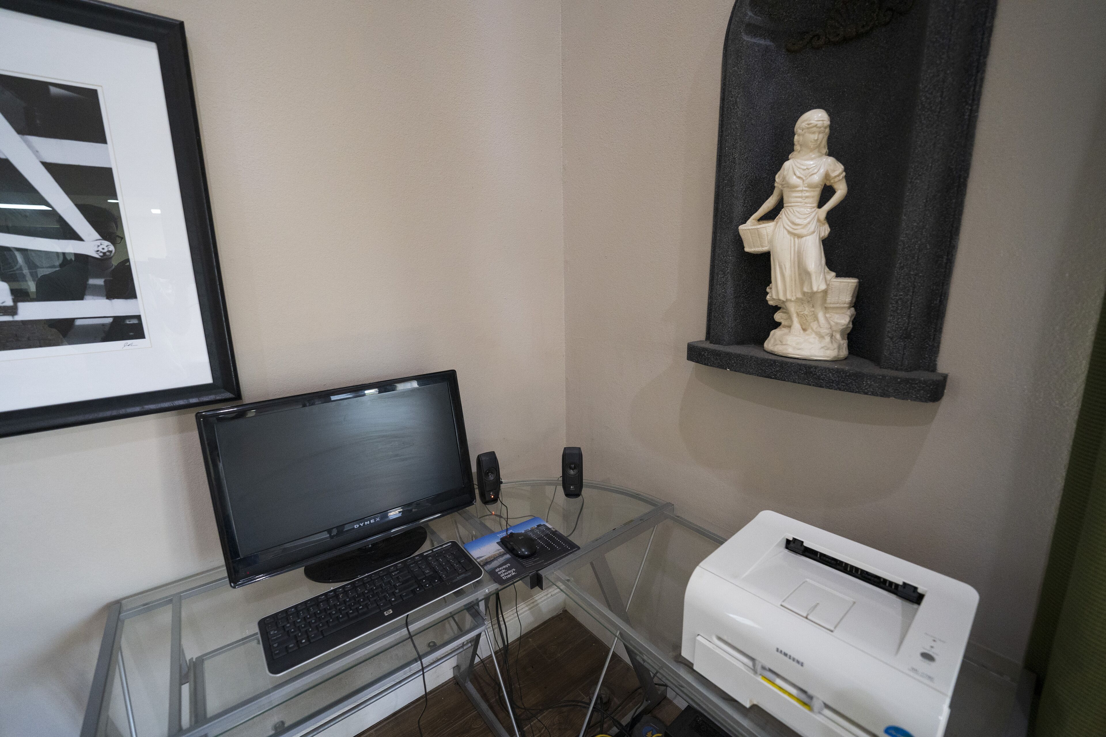 A computer desk with a monitor, keyboard, and printer. There is a framed picture on the wall and a statue on a shelf.