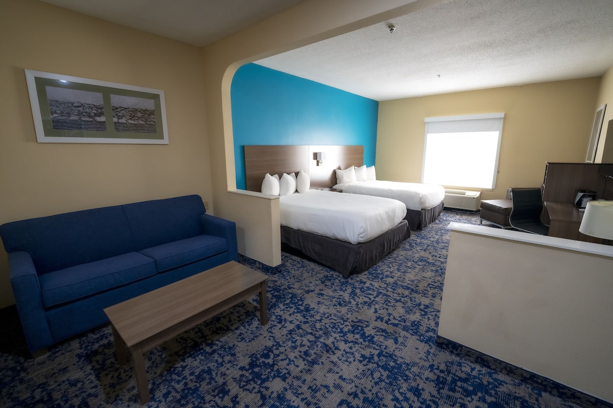 A hotel room with a blue sofa, two beds, a desk, and a large window.