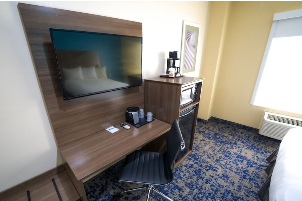 A hotel room with a flat-screen TV mounted on a wooden wall, a desk with a chair, a coffee maker, and a small table with cups.
