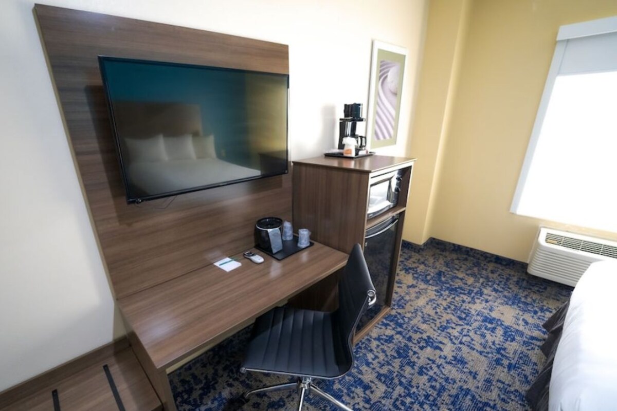 Habitación de hotel con televisor de pantalla plana montado en una pared de madera, un escritorio con una silla, una cafetera y una mesita con tazas.