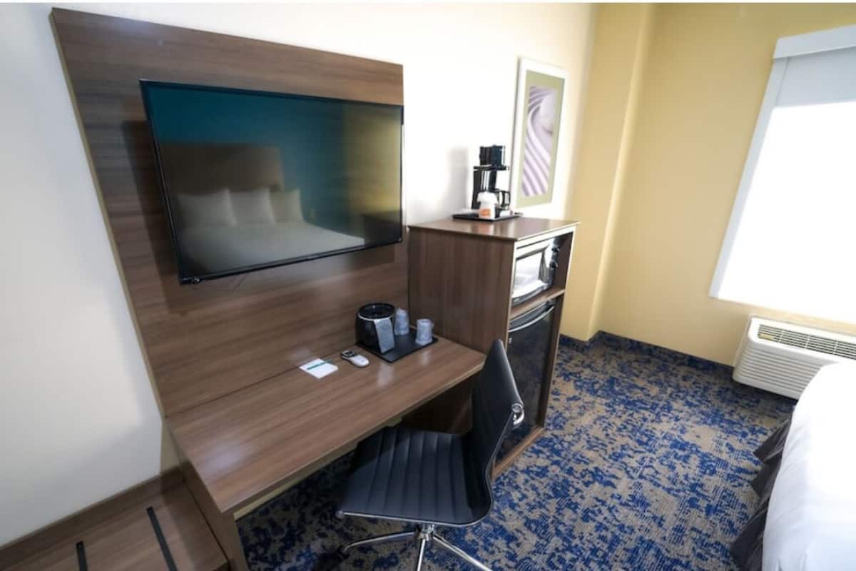 Habitación de hotel con televisor de pantalla plana montado en una pared de madera, un escritorio con una silla, una cafetera y una mesita con tazas.