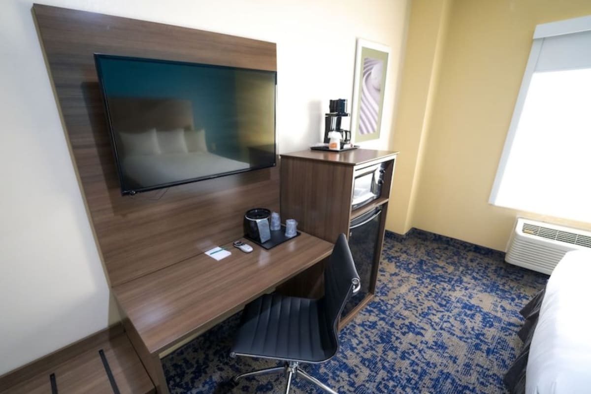 Habitación de hotel con televisor de pantalla plana montado en una pared de madera, un escritorio con una silla, una cafetera y una mesita con tazas.