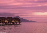 Overwater bungalows with thatched roofs on stilts, connected by walkways, overlooking calm water and distant mountains.