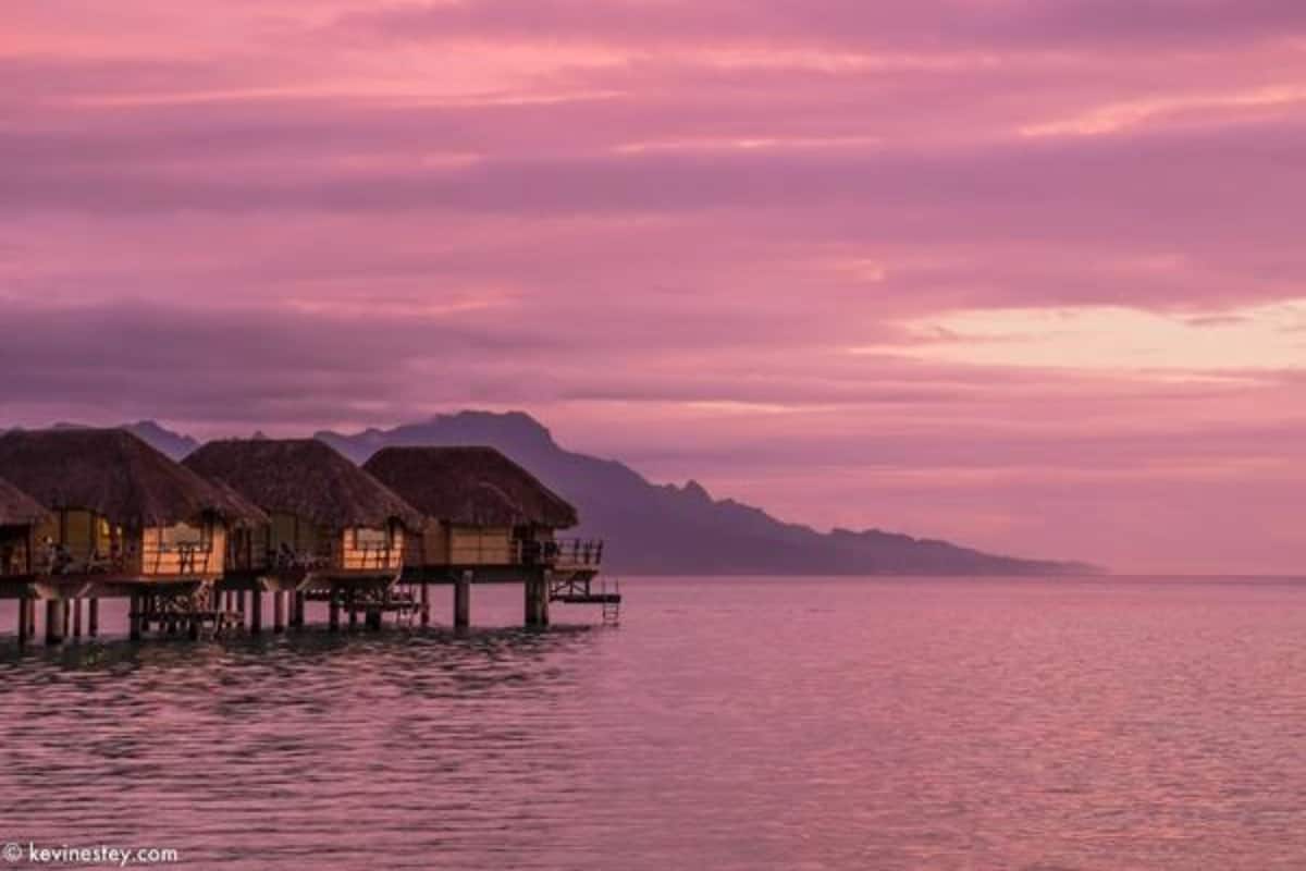 Overwater bungalows with thatched roofs on stilts, connected by walkways, overlooking calm water and distant mountains.