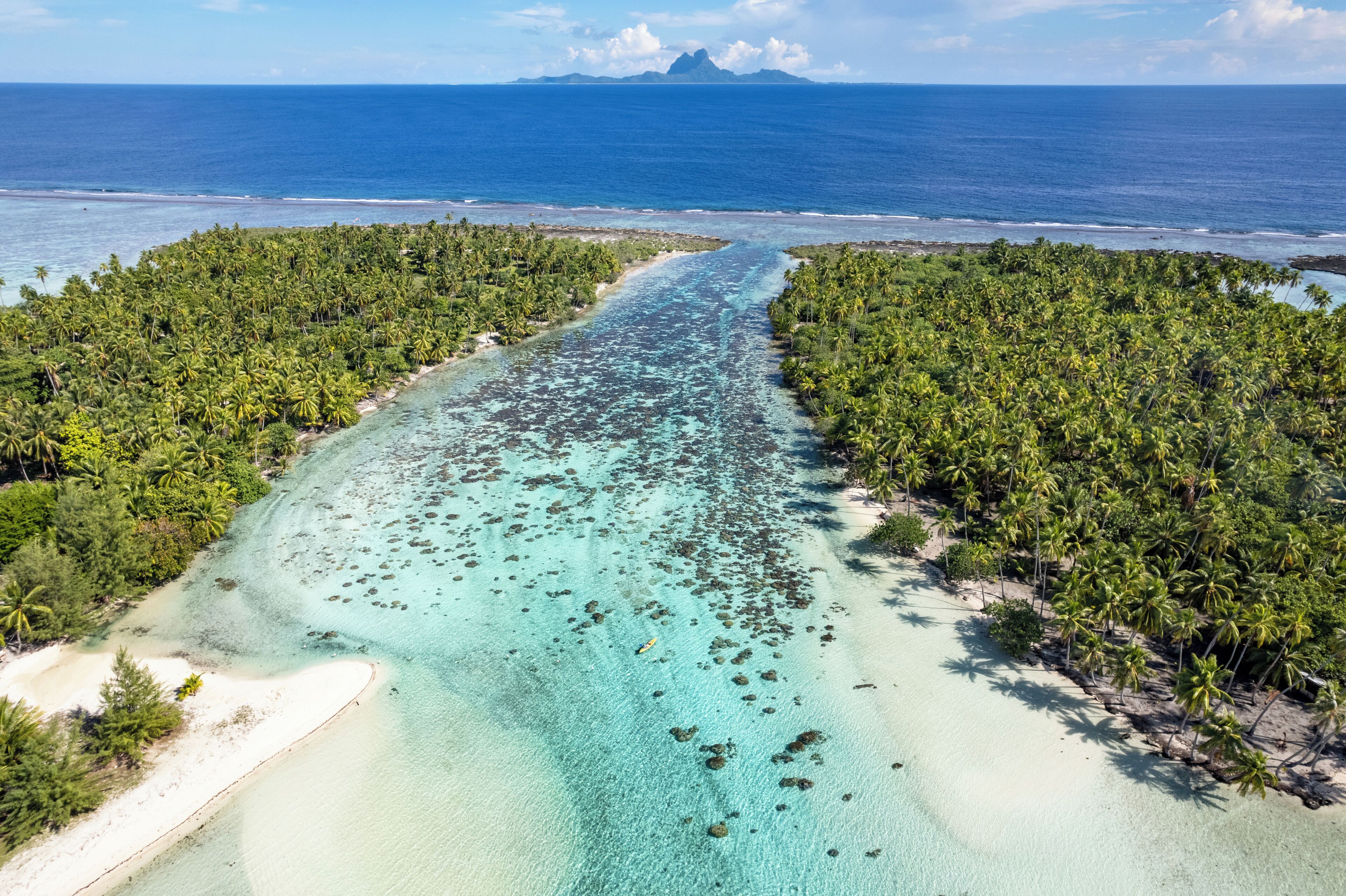 An aerial view of a tropical island with a narrow water channel, dense palm trees, and a clear blue ocean.