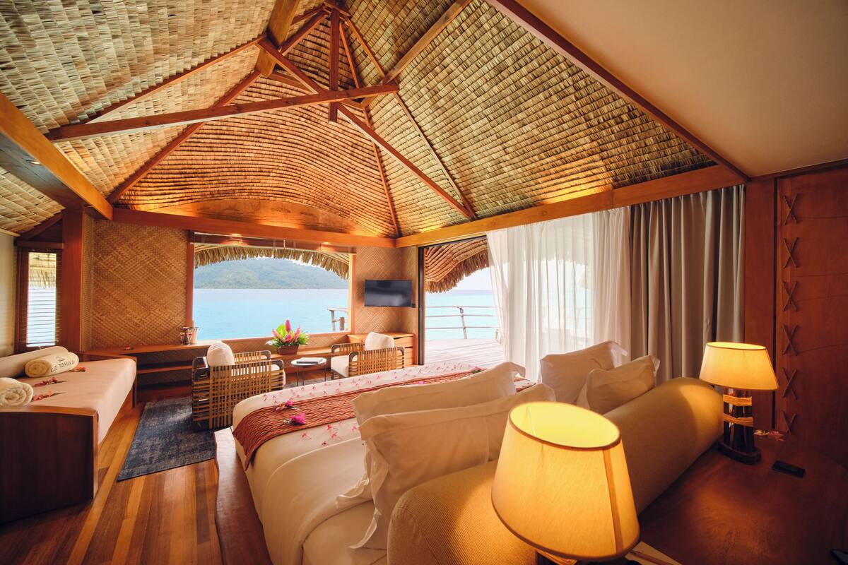 A hotel room with a large bed, a sitting area, a TV, and a view of the sea.
