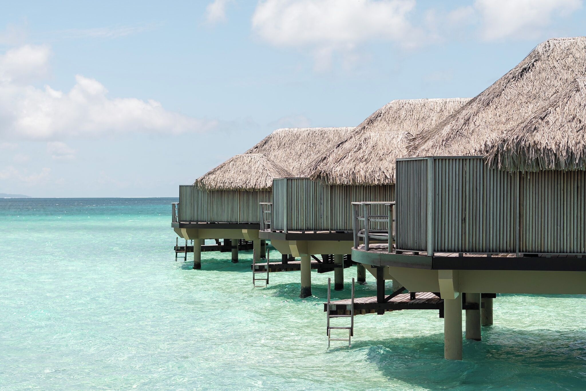 Des bungalows sur pilotis, avec toits de chaume et passerelles en bois, surplombant une eau turquoise et cristalline.