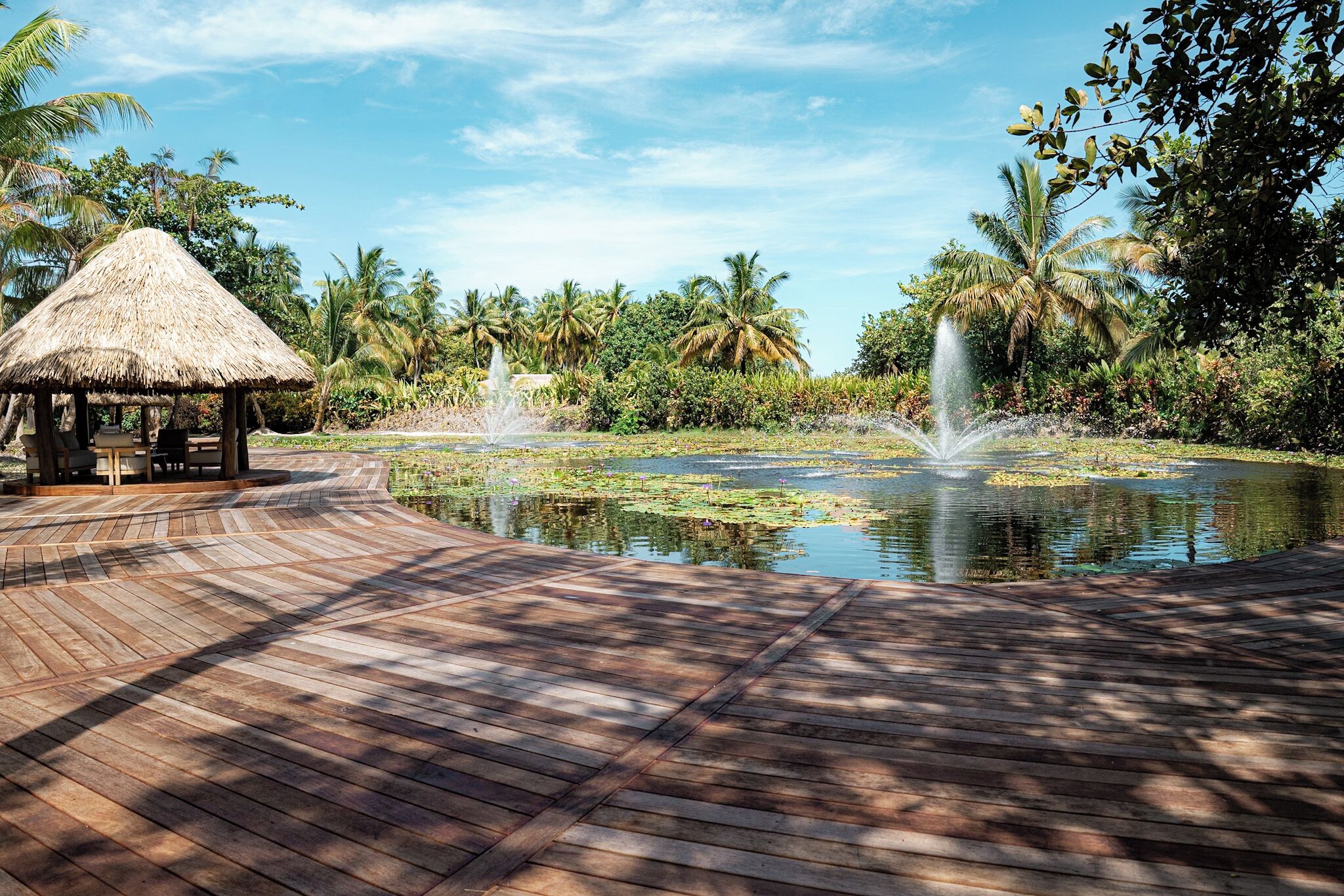 A wooden deck leading to a pond with fountains, surrounded by lush greenery and palm trees.