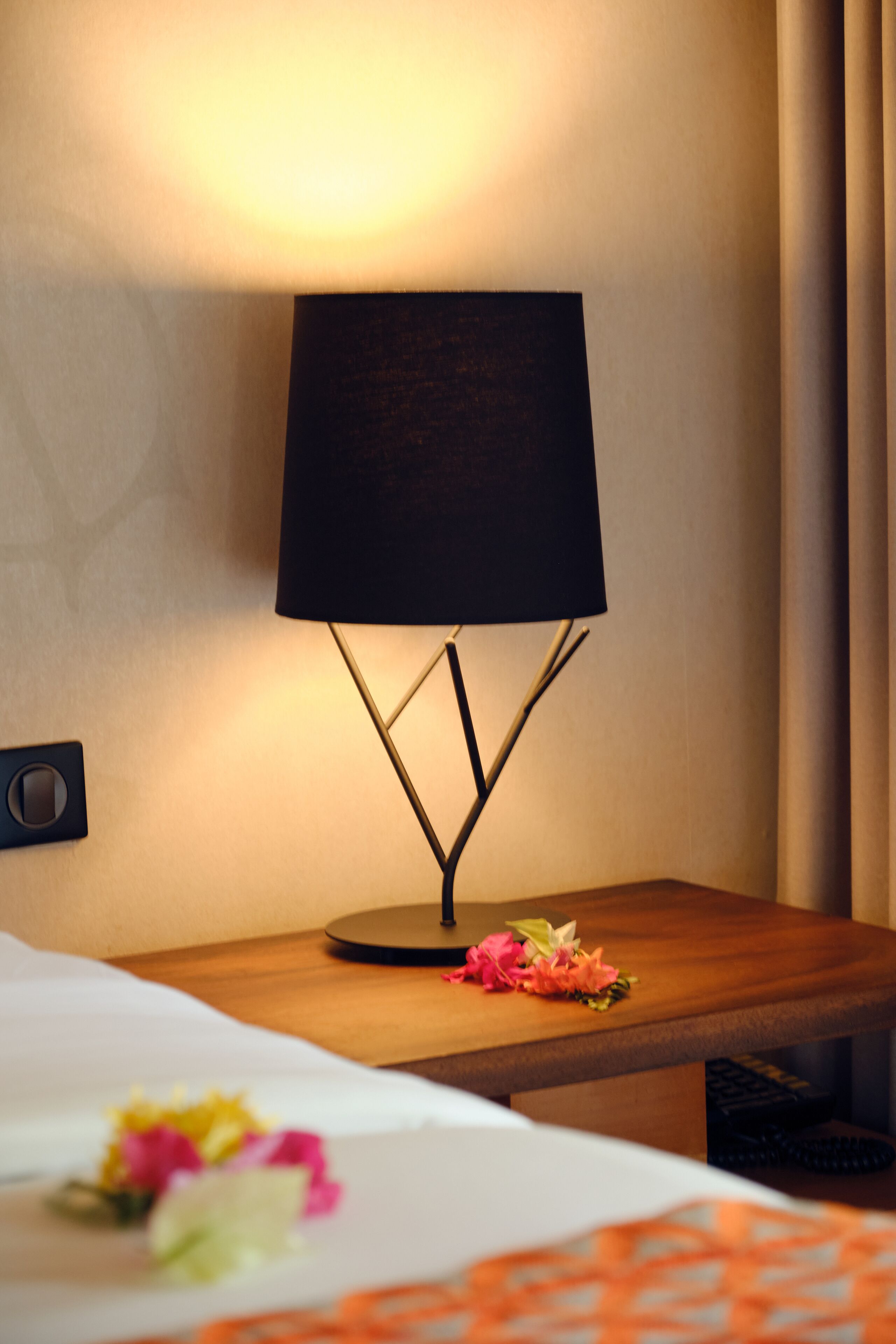 A hotel room with a bedside table, a lamp, and a bed with a floral arrangement.