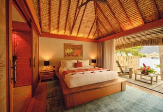 A spacious bedroom with a large bed, wooden headboard, and a view of the sea through a thatched roof.