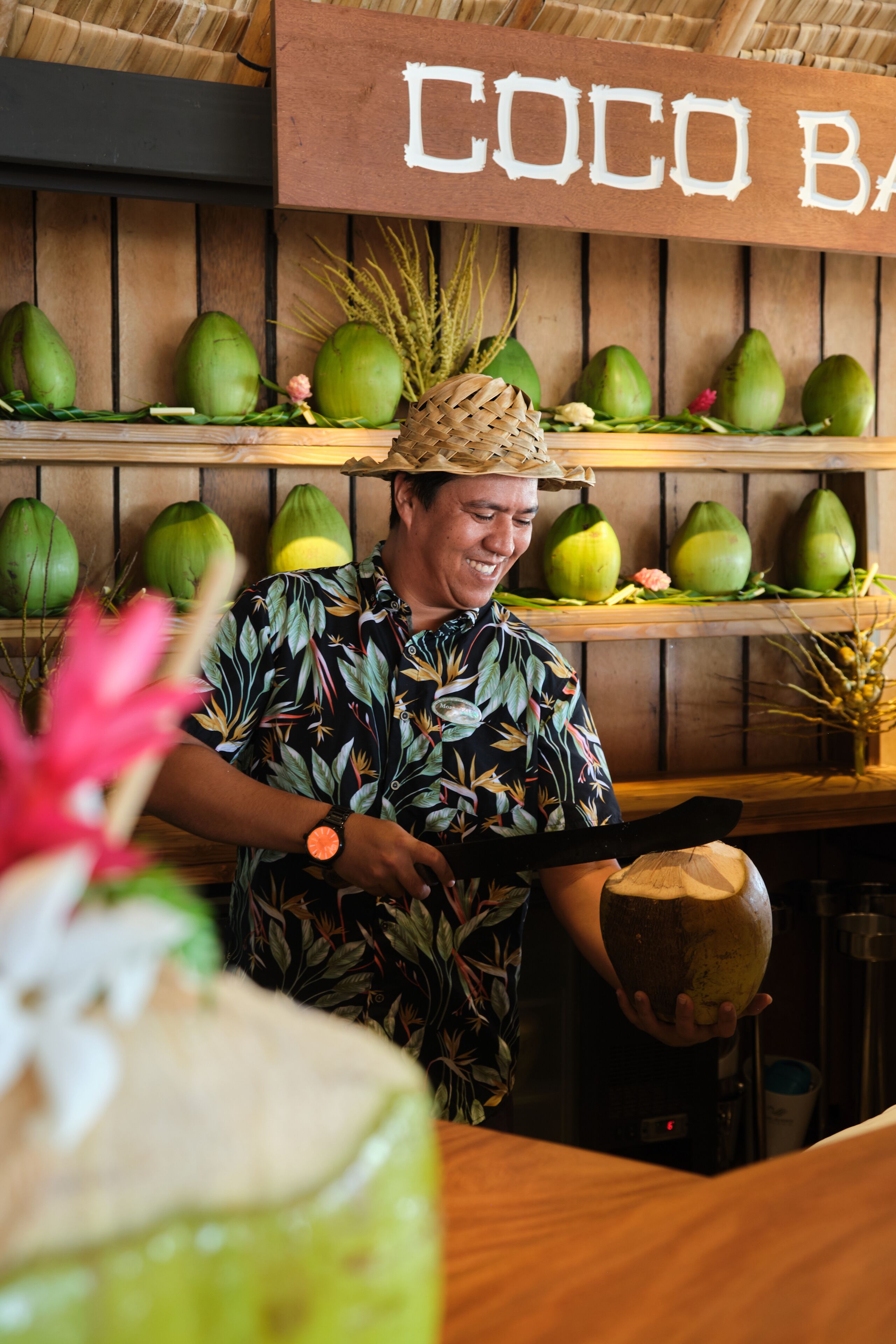 Une personne portant une chemise à motifs tropicaux découpe une noix de coco sur un comptoir, au-dessus duquel est affiché un panneau indiquant « COCO BAR ».