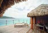 A wooden deck with a lounge chair and a table, overlooking turquoise waters and a distant island.