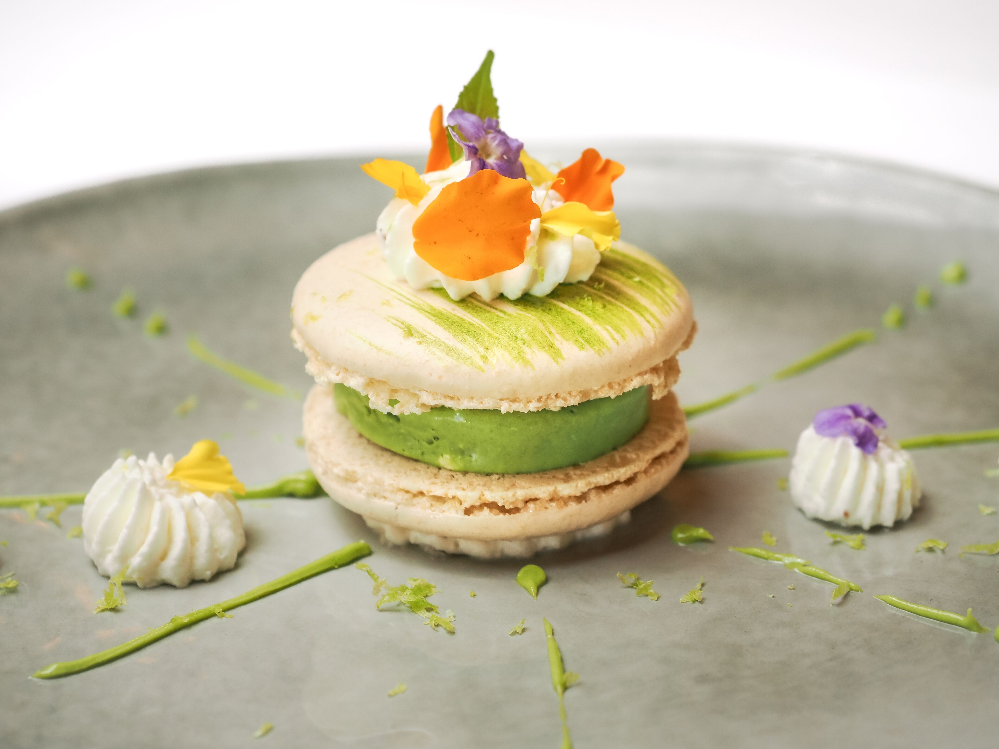 Ein Dessert mit Macarons und grüner Füllung, verziert mit Blumen, auf einem grauen Teller.