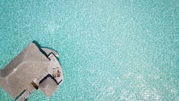 An aerial view of a thatched-roof structure with a wooden deck extending into clear turquoise water.