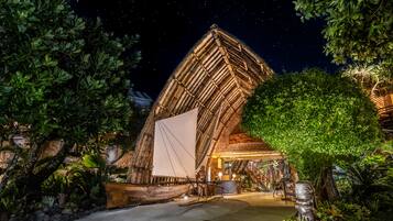 A large, illuminated structure with a thatched roof, surrounded by lush greenery and a clear night sky.