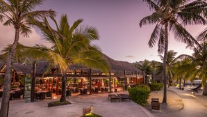 Un restaurant situé en bord de mer, doté d’une terrasse, de palmiers et d’une structure à toit de chaume.