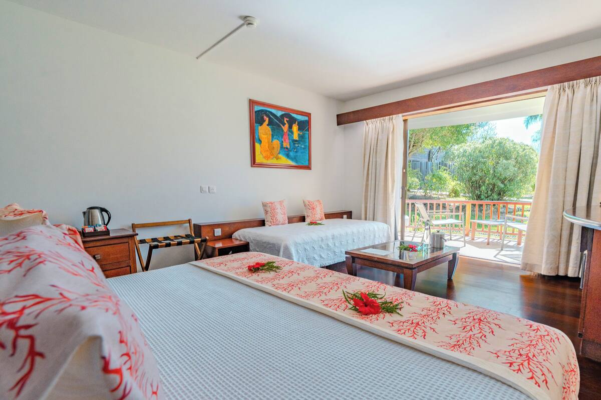 A hotel room with two beds, a painting on the wall, a balcony with a view, and a table with a flower arrangement.