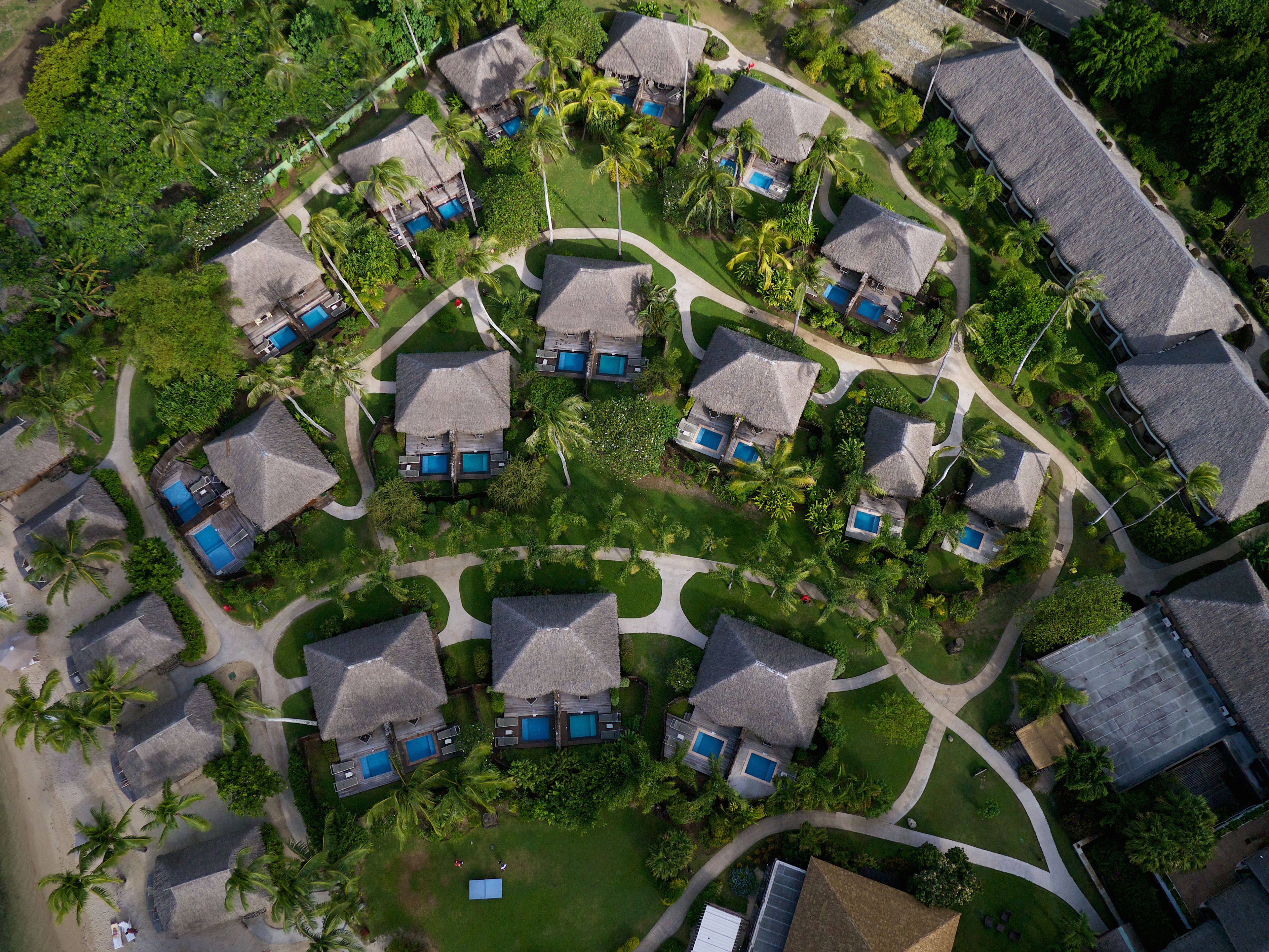 An aerial view of a resort with thatched-roof villas, each with a private pool, surrounded by lush greenery and palm trees.