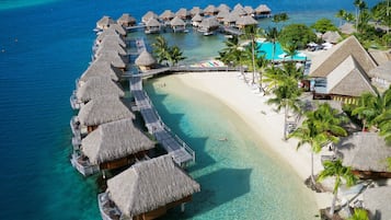 Overwater bungalows connect to a pristine white sandy beach via walkways, surrounded by lush greenery and clear turquoise waters.