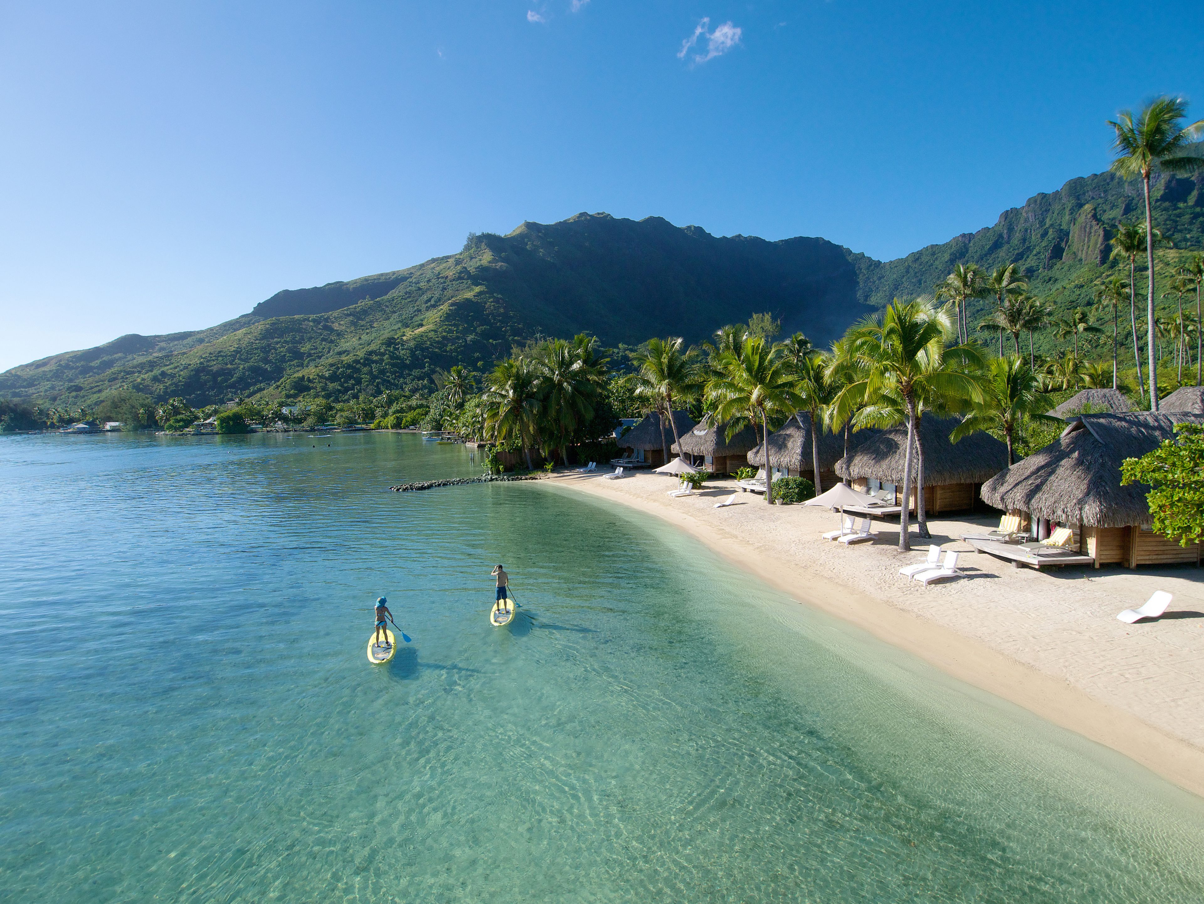 A beach with clear water, palm trees, and thatched-roof huts.