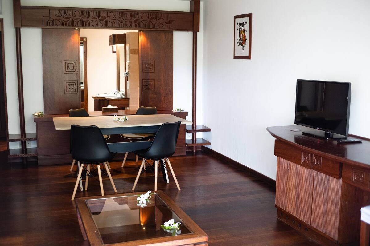 A modern hotel room with a wooden dining table, black chairs, a glass coffee table, a wooden cabinet with a TV, and a decorative wall panel.