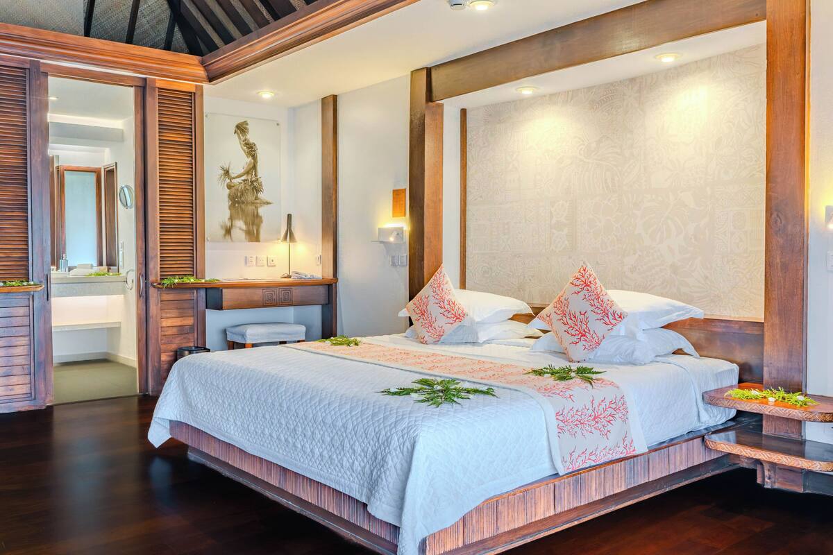 A spacious bedroom with a large bed, a wooden headboard, a nightstand, and a wall-mounted lamp.