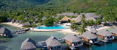 An aerial view of a tropical resort with overwater bungalows, a central swimming pool, and lush greenery in the background.