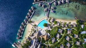 Vue aérienne d’un complexe hôtelier comprenant des bungalows sur pilotis, une piscine centrale et une plage.