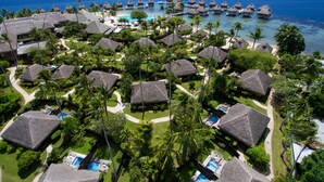 Vue aérienne d’un complexe hôtelier comprenant des bungalows sur pilotis, une piscine et une plage.