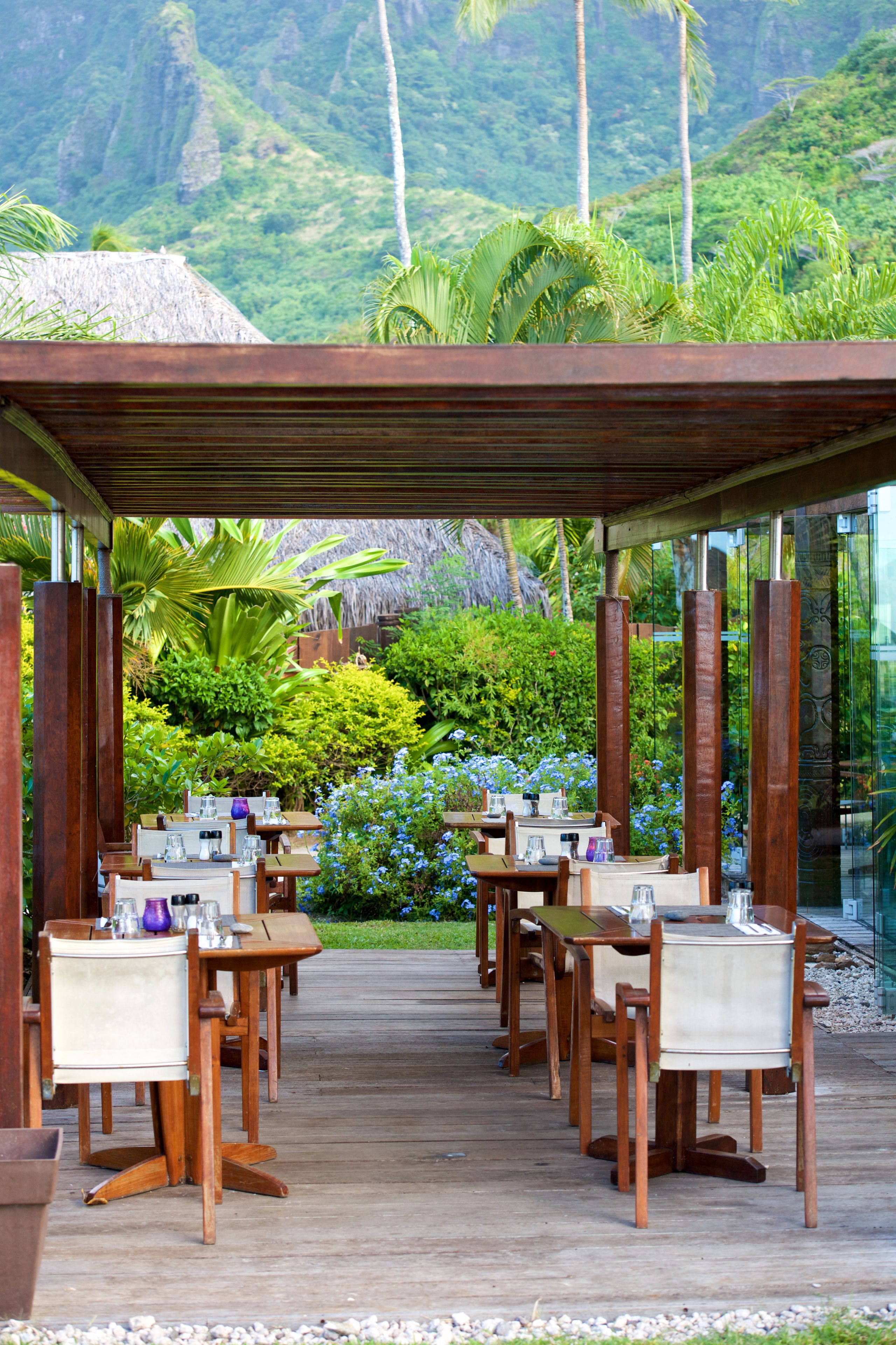 An outdoor dining area with wooden tables and chairs, surrounded by lush greenery and a mountainous backdrop.