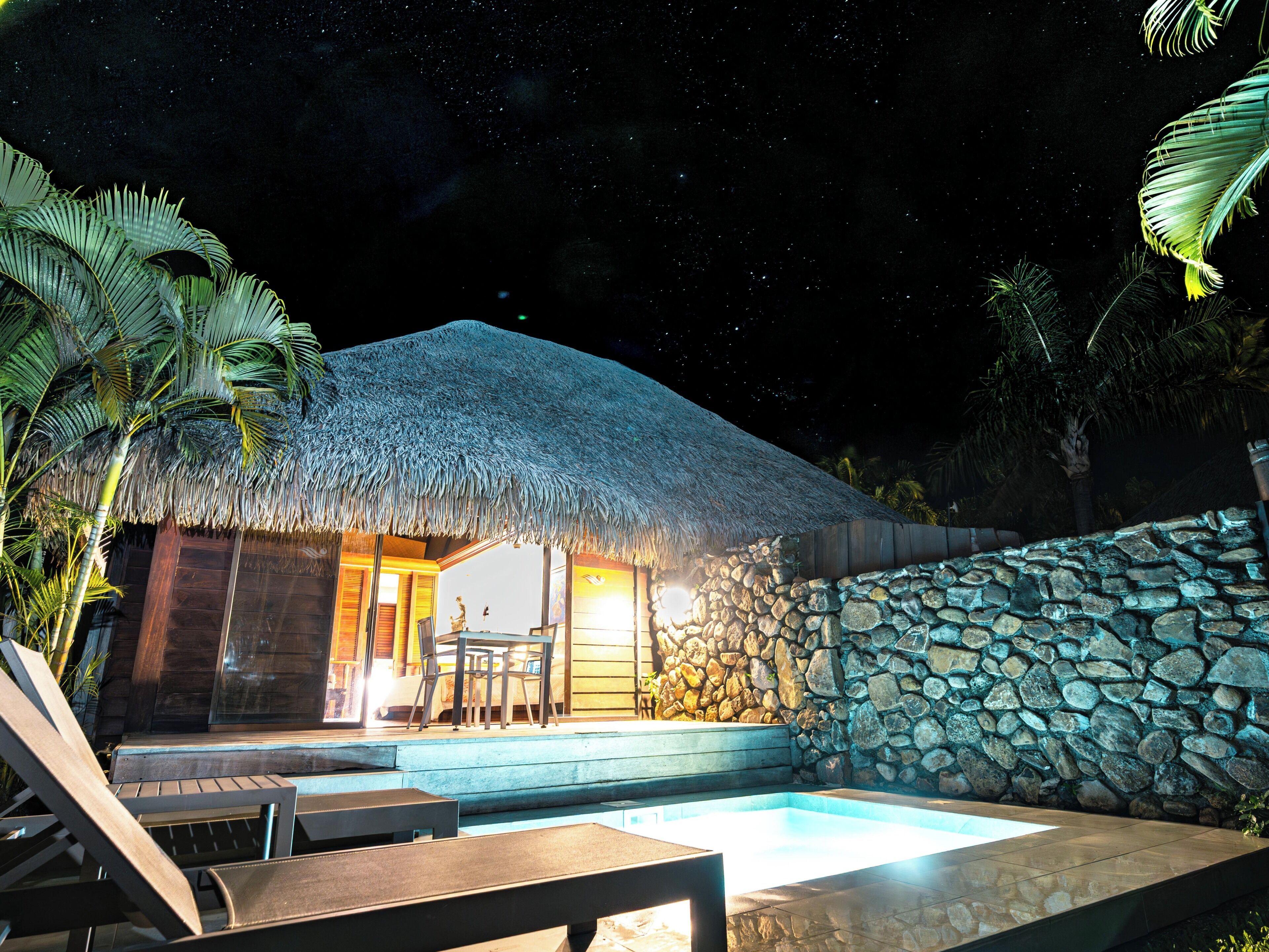 A thatched-roof building with a stone wall, a pool, and lounge chairs.