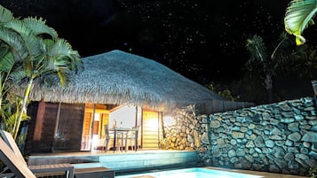 A thatched-roof building with a stone wall, a pool, and lounge chairs.