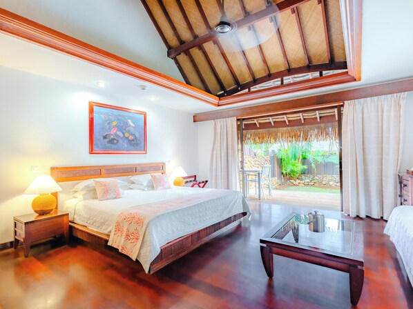 Habitación de hotel con una cama, mesitas de noche, una mesa de centro de vidrio, vista al jardín por la ventana y un ventilador de techo.