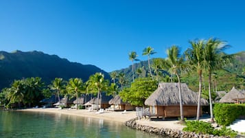 A beach resort with thatched-roof huts, palm trees, and a clear view of mountains in the background.