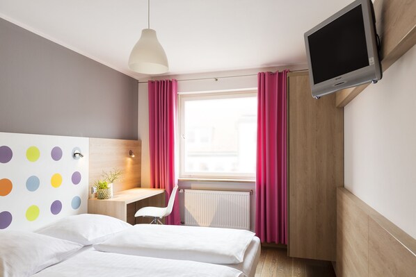 Habitación de hotel con cama, escritorio, silla, televisor y ventana con cortinas.