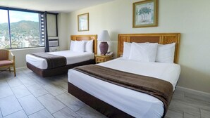 A hotel room with two beds, a chair, a nightstand with a lamp, and a window with a city view. - Waikiki Monarch Hotel (Honolulu)