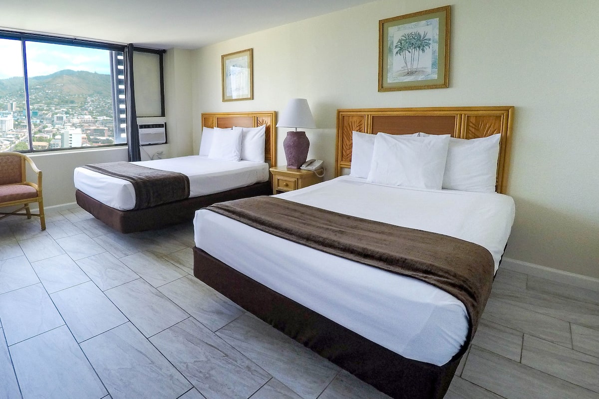 A hotel room with two beds, a chair, a nightstand with a lamp, and a window with a city view.