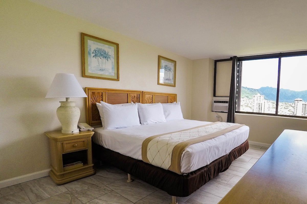A hotel room with a bed, bedside table, lamp, and a window with a city view.