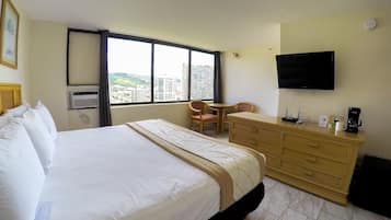 A hotel room with a large bed, a desk, a chair, a dresser, a flat-screen TV, and a window with a city view.