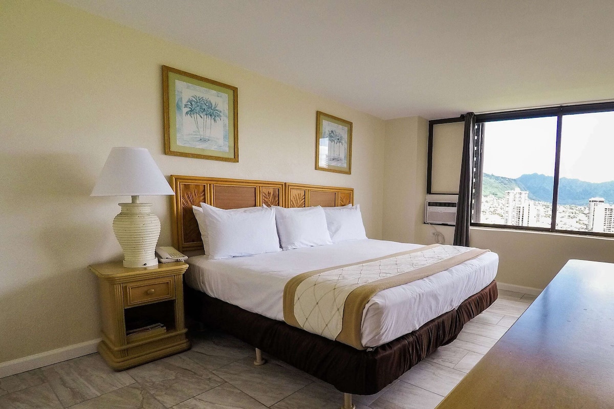 A hotel room with a bed, bedside table, lamp, and a window with a city view.