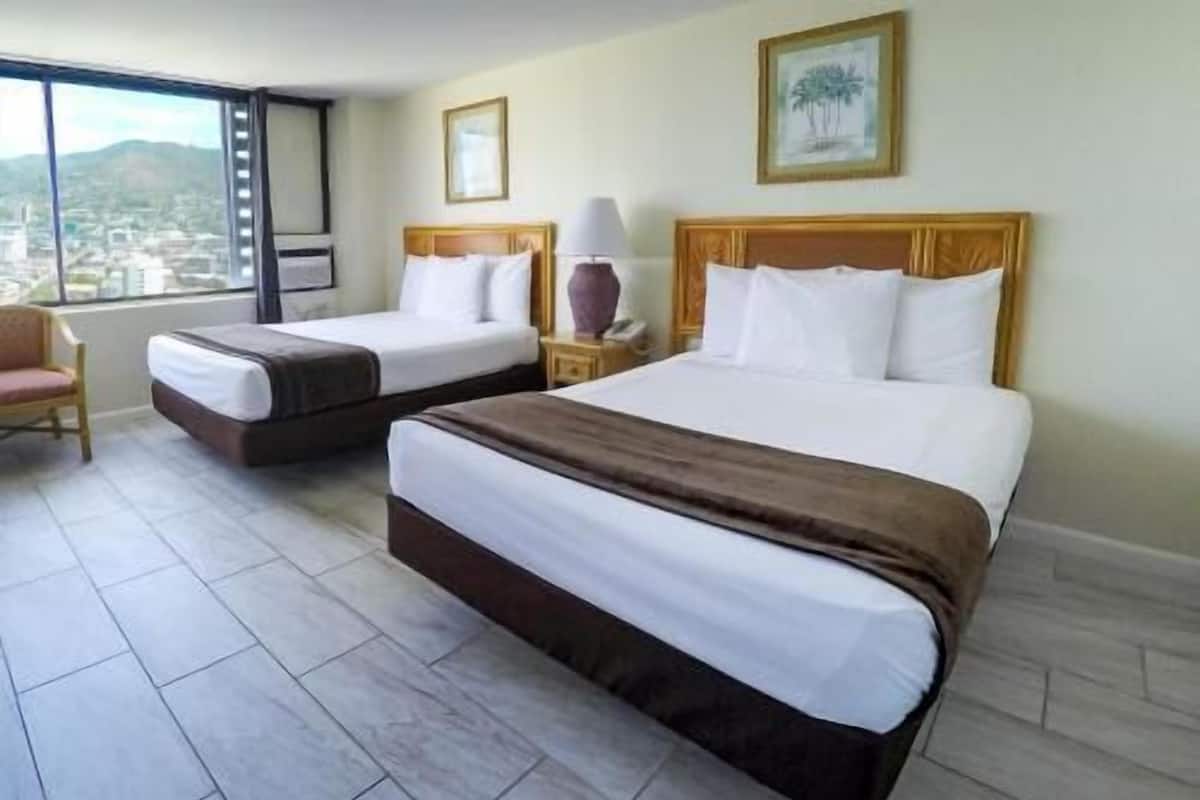 A hotel room with two beds, a chair, a nightstand, a lamp, and a window with a city view.