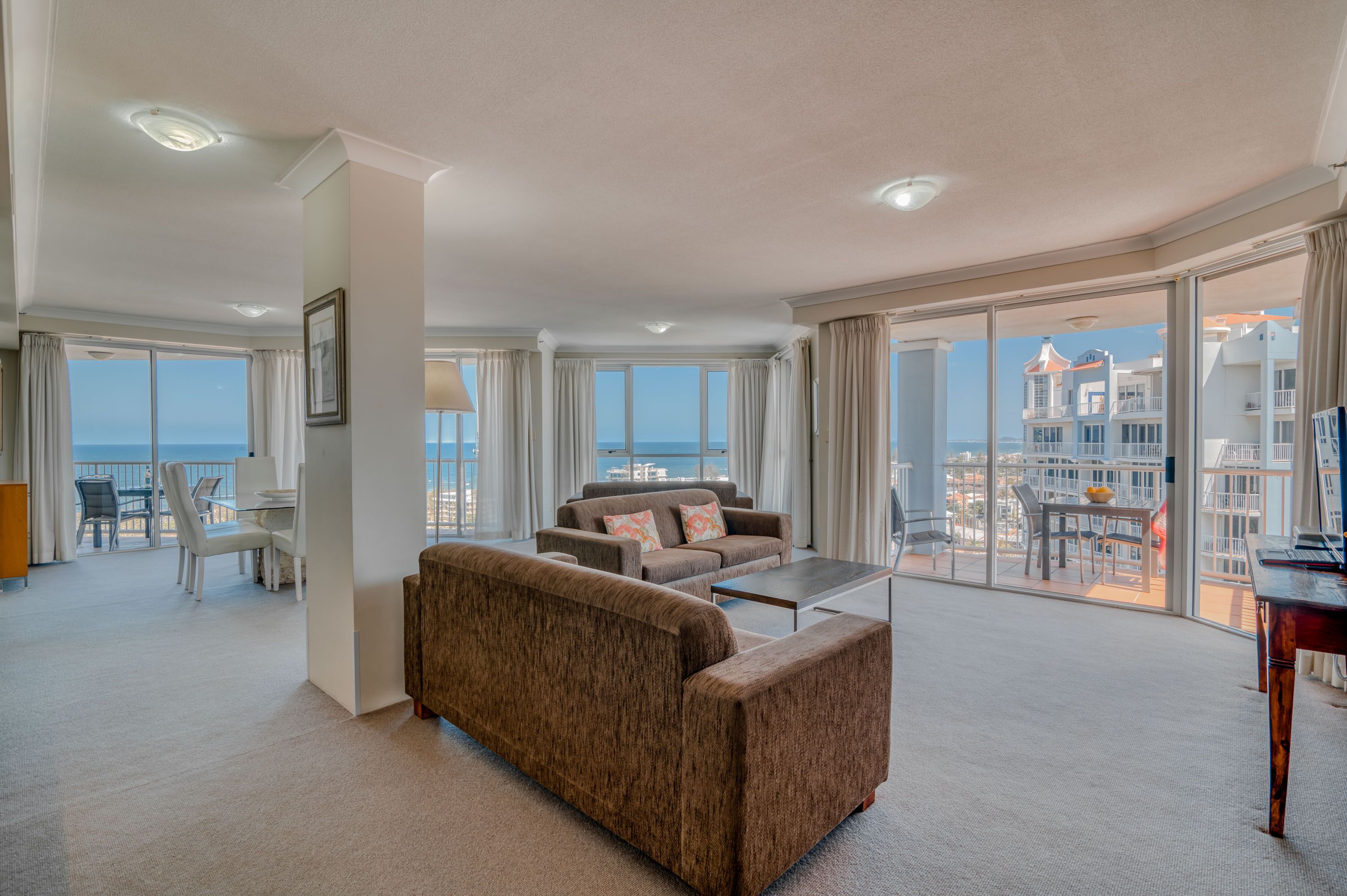 A spacious living room with a large sofa, a coffee table, and a dining area with a table and chairs. The room has large windows offering a view of the cityscape and the sea.