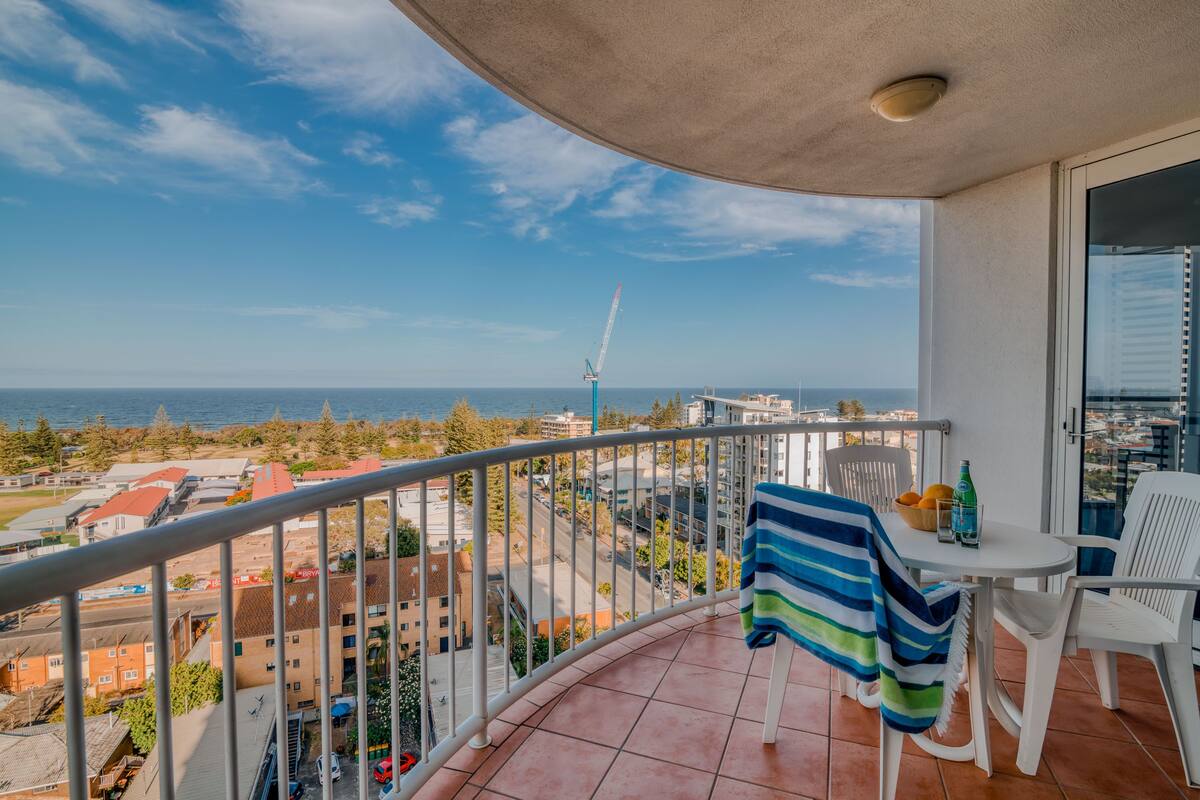 A balcony with a view of the sea and buildings, a table with chairs, and a towel hanging on the railing.